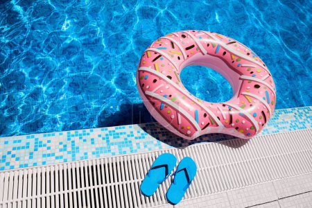 Bague gonflable rose et tongs en caoutchouc bleu par l'eau de piscine extérieure bleue. Détente au bord de la piscine.