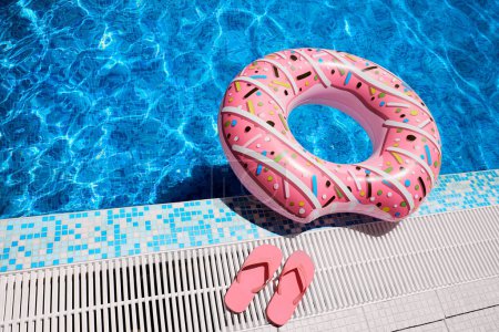 Pink inflatable ring and rubber flip-flops by blue outdoor pool water. Poolside relaxation.