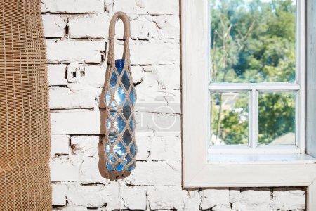 Ein stabiler Seilsack mit einer Wasserflasche, die an einer verwitterten Ziegelwand neben einem sonnenbeschienenen Fenster hängt.