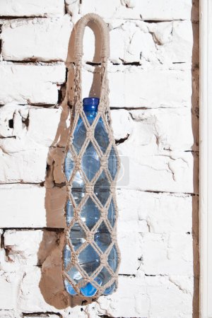 Ein stabiler Seilsack mit einer Wasserflasche, die an einer weißen, verwitterten Ziegelwand hängt.