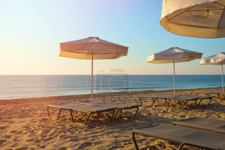 Entspannen am Strand. Ein Paar leere Liegestühle und ein offener Sonnenschirm sitzen an einem Sandstrand, während die Sonne in der Ferne untergeht..