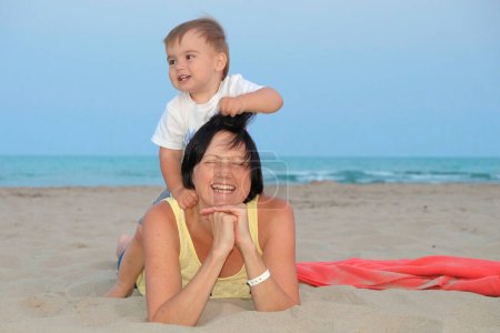 A joyful mother and her son play on a sandy beach with the ocean in the background.
