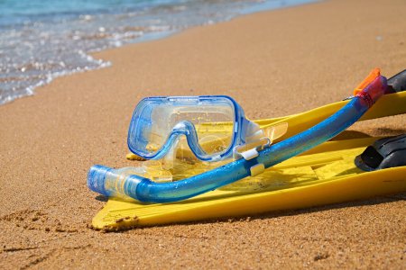 Une paire de palmes jaune vif et un tuba bleu se trouvent sur le sable mou d'une plage tropicale. Les vagues de l'océan turquoise tapissent doucement le rivage en arrière-plan.