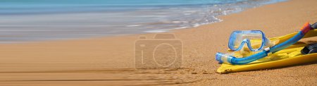 Leuchtend gelbe Schwimmflossen und ein blauer Schnorchel liegen auf dem weichen Sand eines tropischen Strandes. Im Hintergrund plätschern die türkisfarbenen Meereswellen sanft am Ufer entlang.