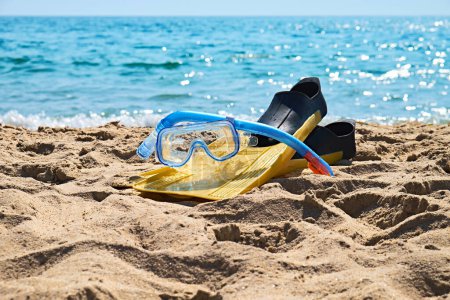 Ein Paar knallgelbe Schwimmflossen und ein blauer Schnorchel liegen auf dem weichen Sand eines tropischen Strandes. Im Hintergrund plätschern die türkisfarbenen Meereswellen sanft am Ufer entlang.