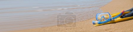 Leuchtend gelbe Schwimmflossen und ein blauer Schnorchel liegen auf dem weichen Sand eines tropischen Strandes. Im Hintergrund plätschern die türkisfarbenen Meereswellen sanft am Ufer entlang.
