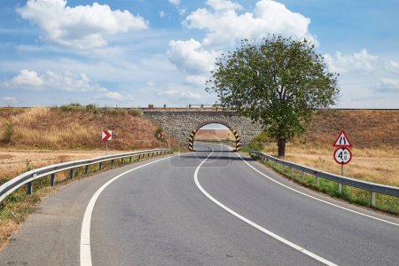 Un camino rural curvo que conduce bajo un puente ferroviario de piedra, flanqueado por barandillas y señales de tráfico. Un árbol solitario está a la derecha, con un cielo azul claro y nubes dispersas sobre la cabeza.