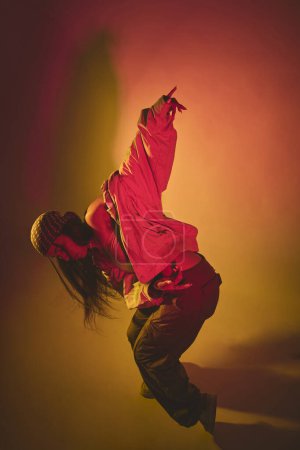 A woman in a white shirt and brown pants is dancing in front of a red background. The image has a vibrant and energetic mood, with the womans movements