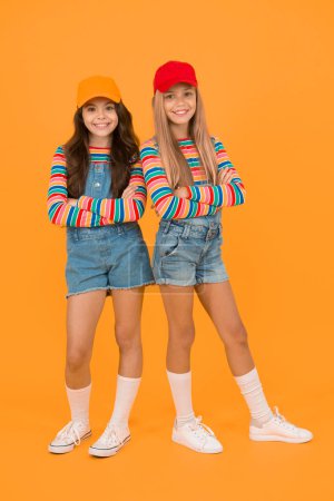 Fashion inspired by the sneaker culture. Happy kids keeping arms crossed with fashion look. Fashion small girls in casual wear smiling on yellow background. Streetwear fashion for little children.