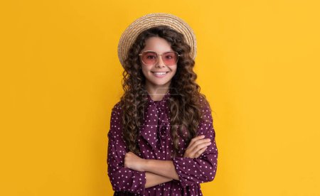 smiling child in straw hat and sunglasses with long brunette curly hair on yellow background.