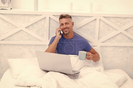 Hablando por teléfono. El tipo feliz habla por celular usando el portátil en la cama. Charlando en línea. Café de la mañana en la cama.