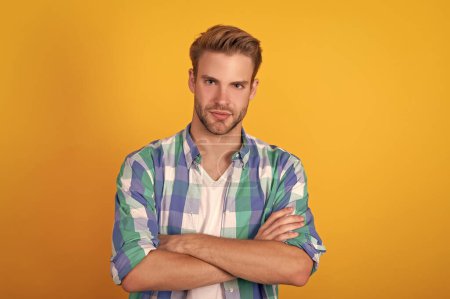 Serious handsome young man portrait keeping arms crossed in checked shirt yellow background, confident.