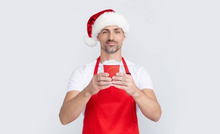 mature christmas man in red santa hat and apron holding coffee cup.