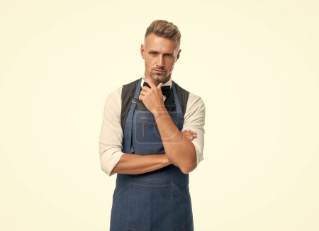 Serious guy in barista apron and bow stroking chin isolated on white.