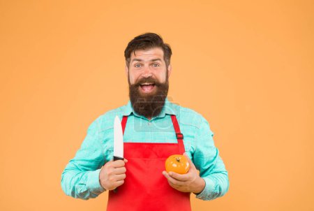 Bearded man in red apron with yellow tomato. cooking healthy and fresh food. Handsome male chef is working. mature cook cut vegetable. guy in uniform on kitchen. vitamin dieting.