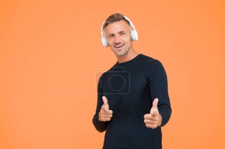 Handsome cheerful man listen music in stylish headphones on yellow background pointing finger, having fun.