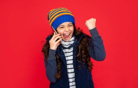 glad kid talking on phone in hat on red background. teen girl in down vest with smartphone. portrait of child greeting while phone conversation. communication. happy winter holidays.