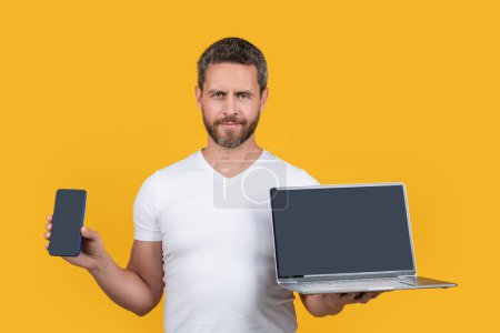 Kerl zeigt Bildschirm mit Kopierraum. Kerl zeigt Handybildschirm isoliert auf gelbem Hintergrund. Mann zeigt Telefon und Laptop im Studio. Mann zeigt Bildschirm von Laptop und Handy.