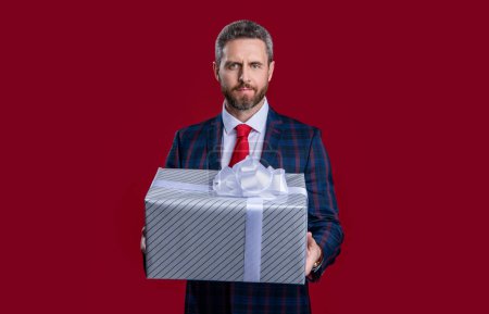 Hombre de negocios presente en el estudio. foto del hombre de negocios con presente. Hombre de negocios sosteniendo la caja de regalo en traje. hombre de negocios con presente aislado sobre fondo rojo.
