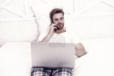 Photo for Unhaven guy in sleepwear relax in bed talking on mobile phone computer technology, conversation. - Royalty Free Image