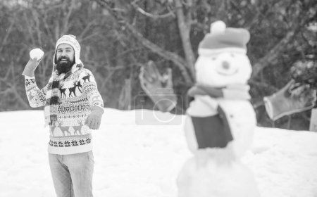 Leisure on fresh air. Snowman and cheerful bearded hipster knitted hat and warm gloves play with snow outdoors. Have fun winter day. Let it snow. Christmas holidays. Active lifestyle. Snow games.