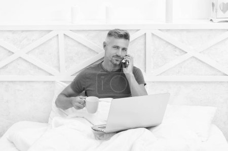 Foto de Hombre feliz hablar en el teléfono celular beber café y el uso de la computadora portátil en la cama por la mañana, móvil. - Imagen libre de derechos