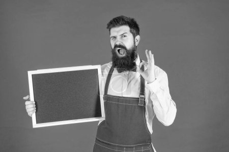 Photo for Happy winking man in barber apron hold school blackboard showing OK sing ring gesture red background copy space, enrollment. - Royalty Free Image