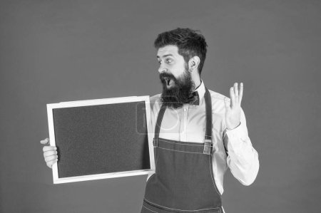Photo for Surprised bearded man in barber apron hold school blackboard red background copy space, enrollment. - Royalty Free Image
