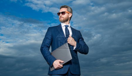 photo of confident businessman with laptop. businessman with laptop on sky background. businessman with laptop outdoor. businessman with laptop wearing suit.