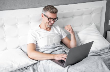 Téléchargez les photos : Heureux homme mûr dans des lunettes de travail sur ordinateur portable dans le lit. - en image libre de droit