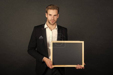 Photo for Serious guy in formal suit holding empty school blackboard copy space dark background, teacher. - Royalty Free Image