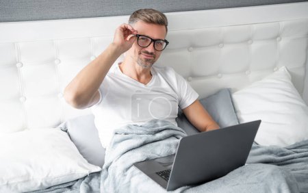 Téléchargez les photos : Mature man in glasses check email on laptop in bed. - en image libre de droit