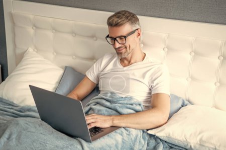 Téléchargez les photos : Mature guy dans lunettes de travail sur ordinateur portable dans lit. - en image libre de droit