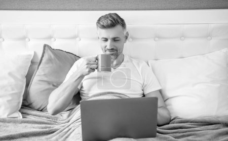 smiling mature man working on laptop in bed with coffee.