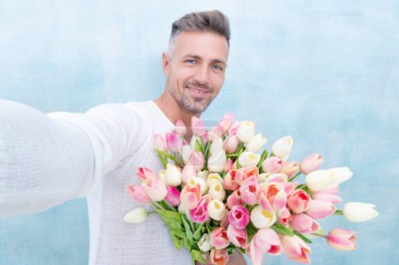 man with valentines tulips taking selfie photo isolated on blue background. photo of man with valentines tulips. man with valentines tulips. man with valentines tulips in studio.