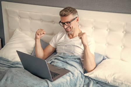 Photo for Successful mature man in glasses working on laptop in bed. - Royalty Free Image