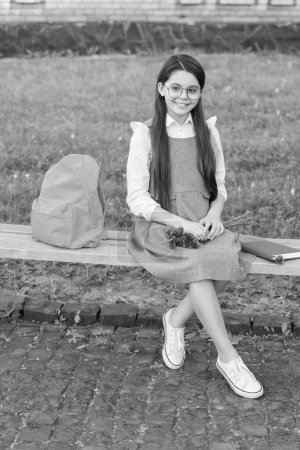 Téléchargez les photos : Enfant heureux dans des lunettes se détendre sur le banc de l'école avec des fleurs et sac à dos. Journée des femmes. - en image libre de droit