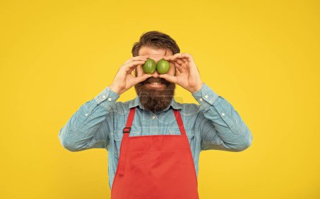 Foto de Happy guy in apron holding fresh limes on eyes yellow background, fruit seller. - Imagen libre de derechos