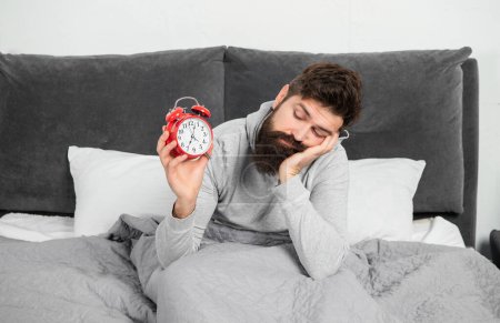 Drowsy man looking at alarm clock being in bed in morning, sleep loss.
