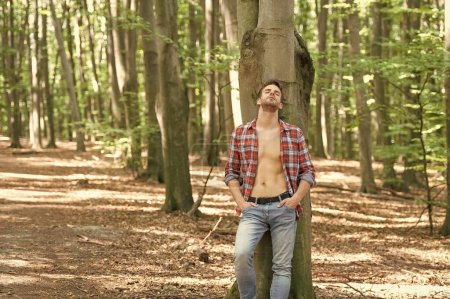 Téléchargez les photos : Homme Lumbersexual avec les yeux fermés en chemise bûcheron déboutonnée debout à l'arrière-plan de la forêt d'arbres, espace de copie. - en image libre de droit