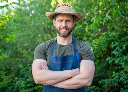 Happy gardener man smiling in gardening apron and farmers hat keeping arms crossed in garden outdoors.