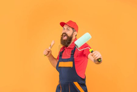 angry bearded man in work clothes hold paint roller and brush on yellow background.