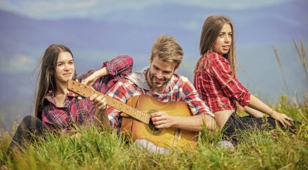 Photo for Friends hiking with music. Nothing but friends and guitar. People relaxing on mountain top while handsome man playing guitar. Hiking entertainment. Peaceful place. Melody of nature. Hiking tradition. - Royalty Free Image