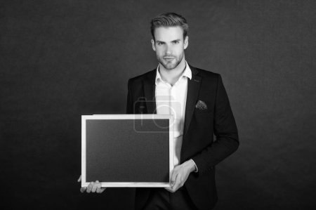 Photo for Serious guy in formal suit holding empty school blackboard copy space dark background, teacher. - Royalty Free Image
