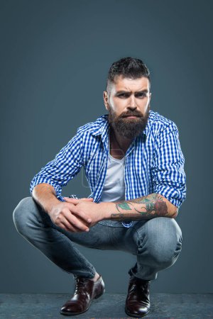 bearded guy wear checkered shirt sitting on grey background.