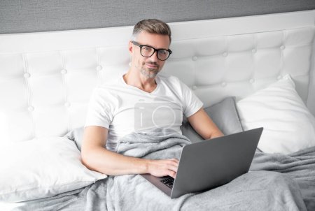 Téléchargez les photos : Mature man virtual assistant in glasses working on laptop in bed. - en image libre de droit