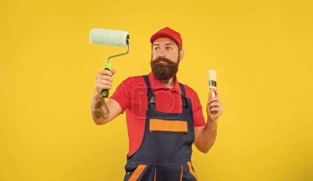 puzzled bearded man in work clothes hold paint roller and brush on yellow background.