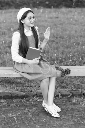 Téléchargez les photos : Enfant heureux avec geste bonjour relax tenant livre sur le banc du parc. - en image libre de droit