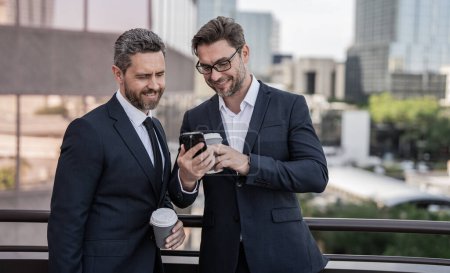 photo of smiling business executive check phone. business executive check phone in the street. business executive check phone outdoor. business executive in suit check phone holding coffee.