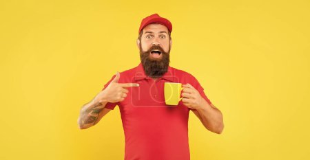Téléchargez les photos : Surpris homme barbu en chapeau rouge décontracté et tshirt pointant du doigt sur fond jaune tasse, thé. - en image libre de droit
